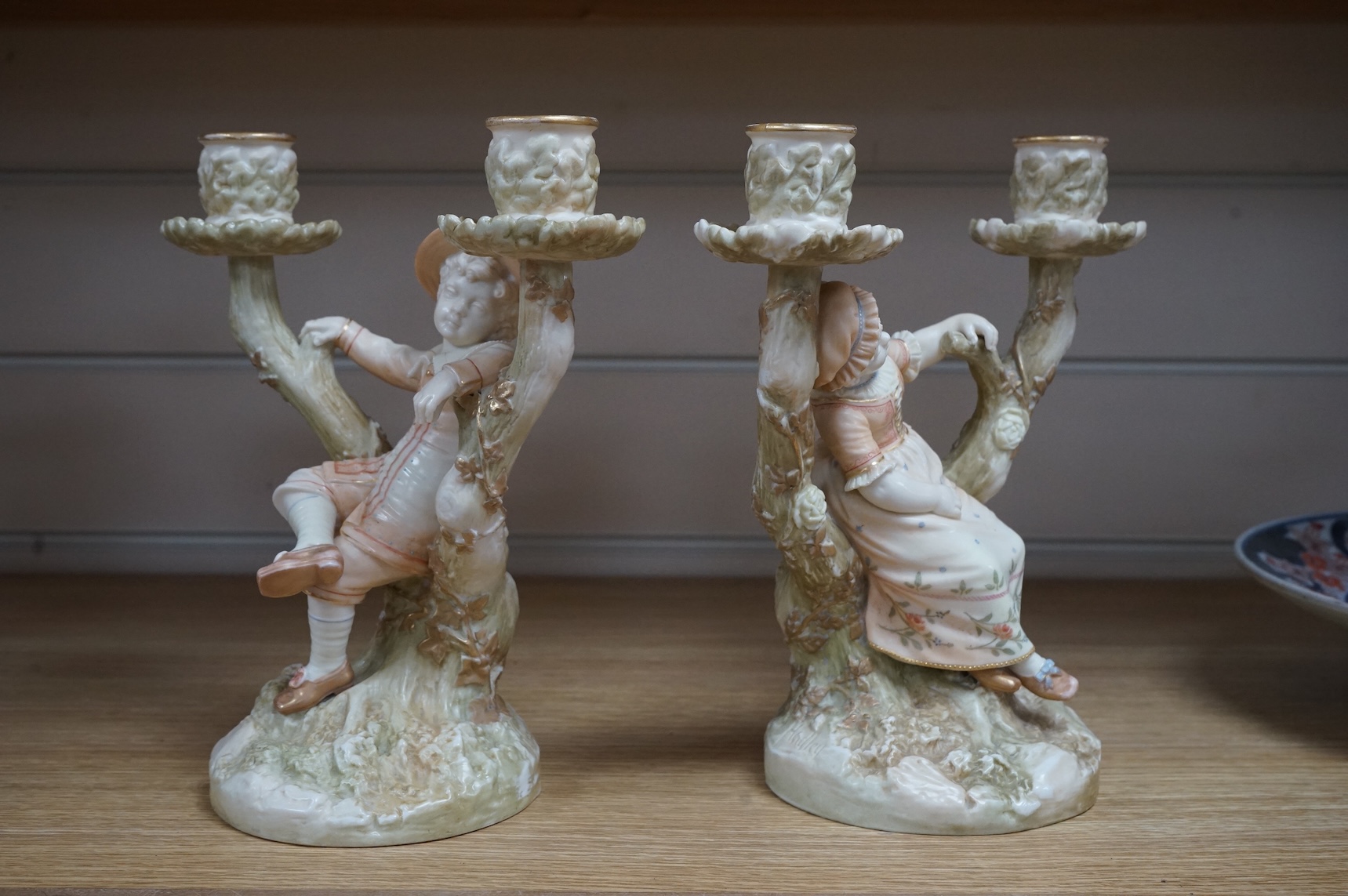 A pair of late 19th century Royal Worcester Kate Greenaway style figural two branch candelabra, modelled by James Hadley 22.5cm high